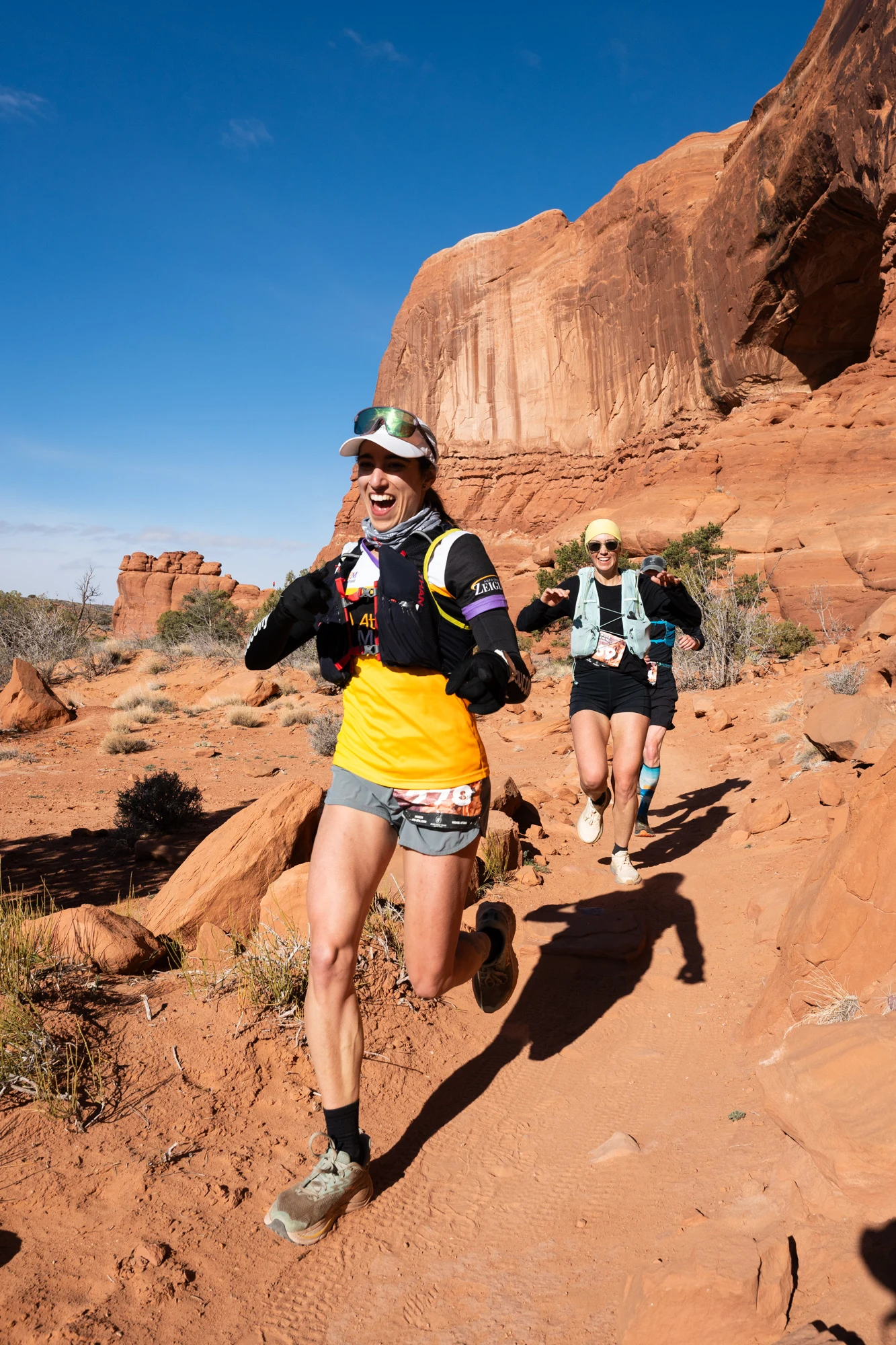 A smiling trail runner leads two others through a red-rock canyon.