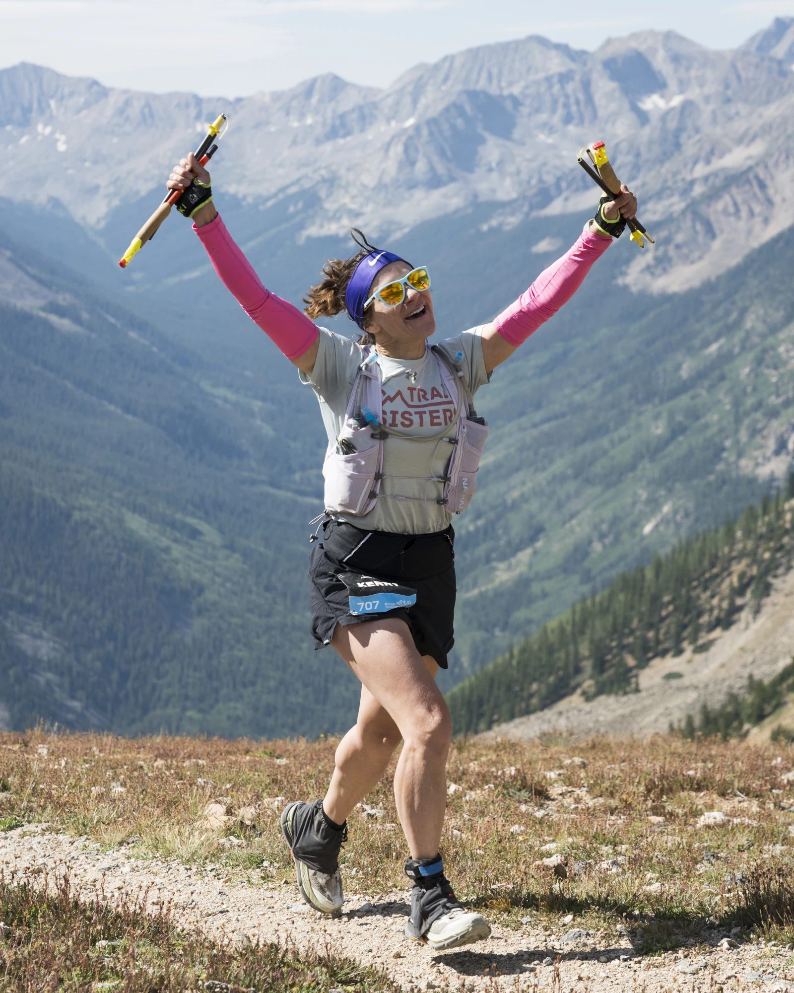 A runner raises both arms on a high-alpine trail with mountain slopes behind her.