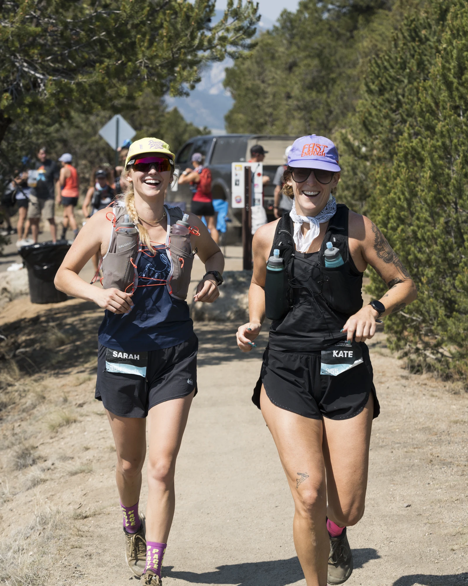 Two smiling trail runners run side by side near a TransRockies aid station.