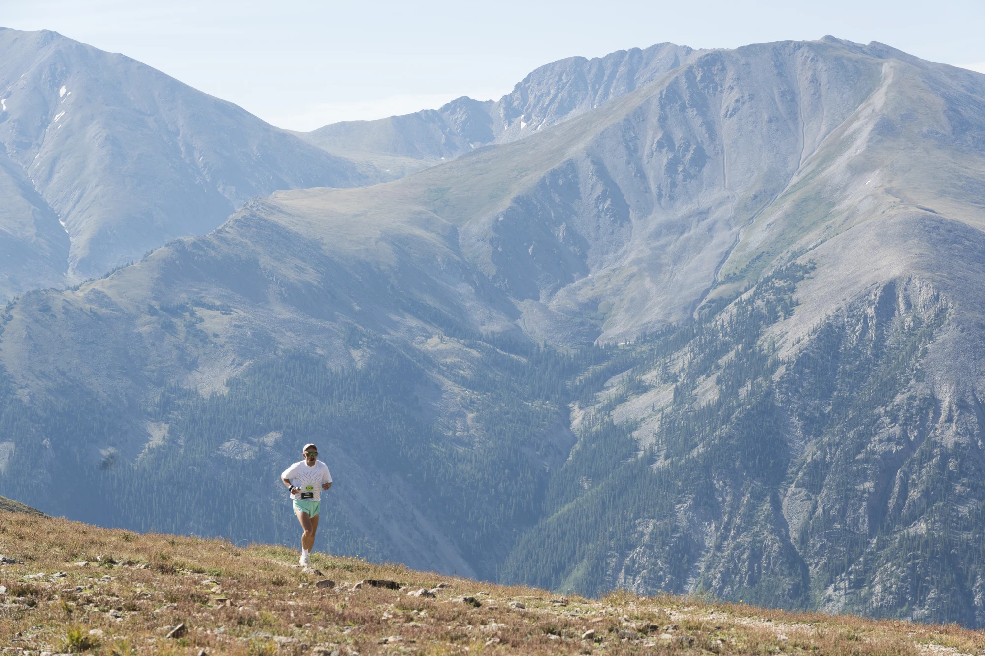 A lone trail runner crosses a high-alpine slope with mountain ridges behind him.