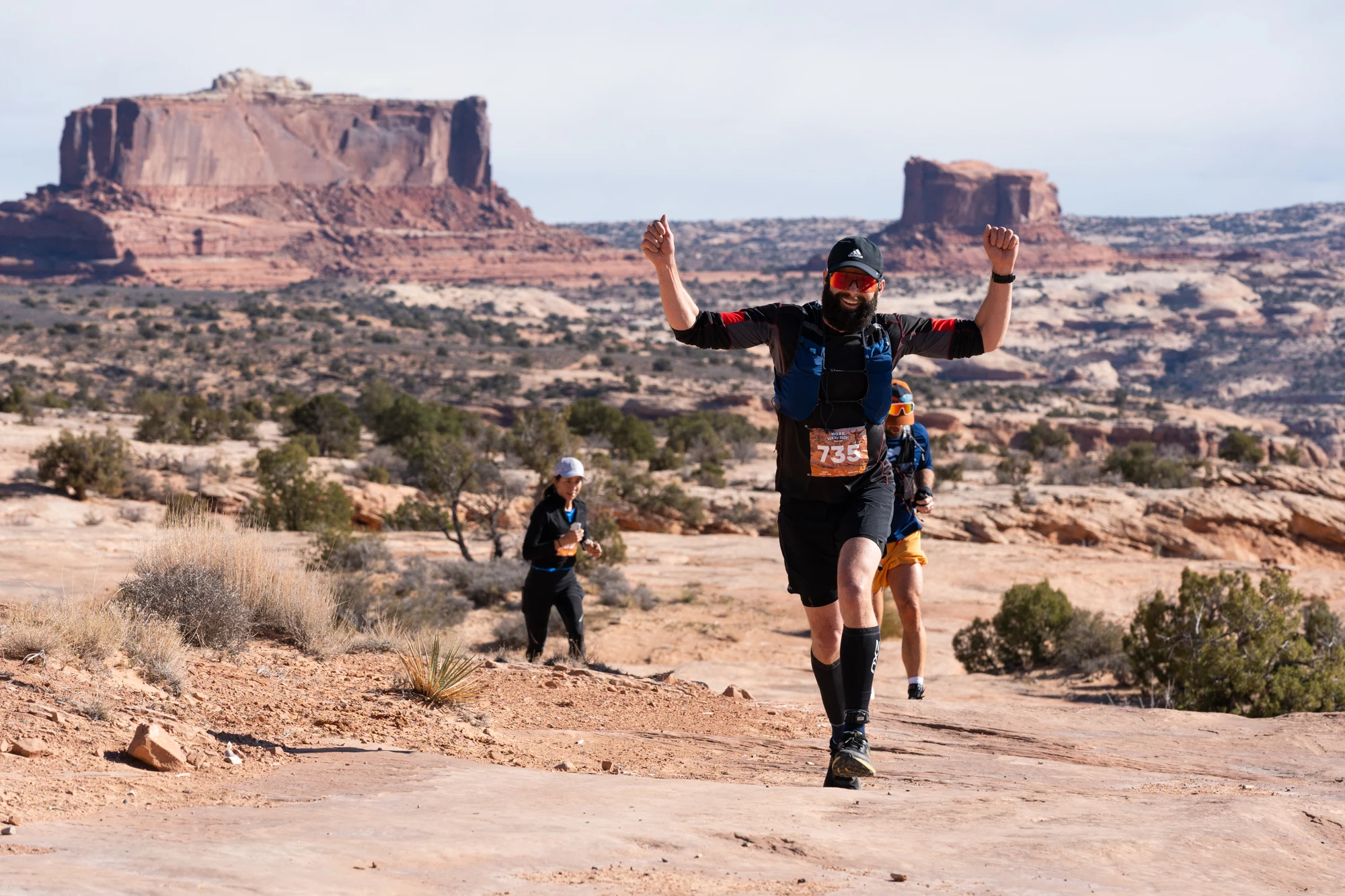 A trail runner raises both arms on desert slickrock beneath red-rock mesas.