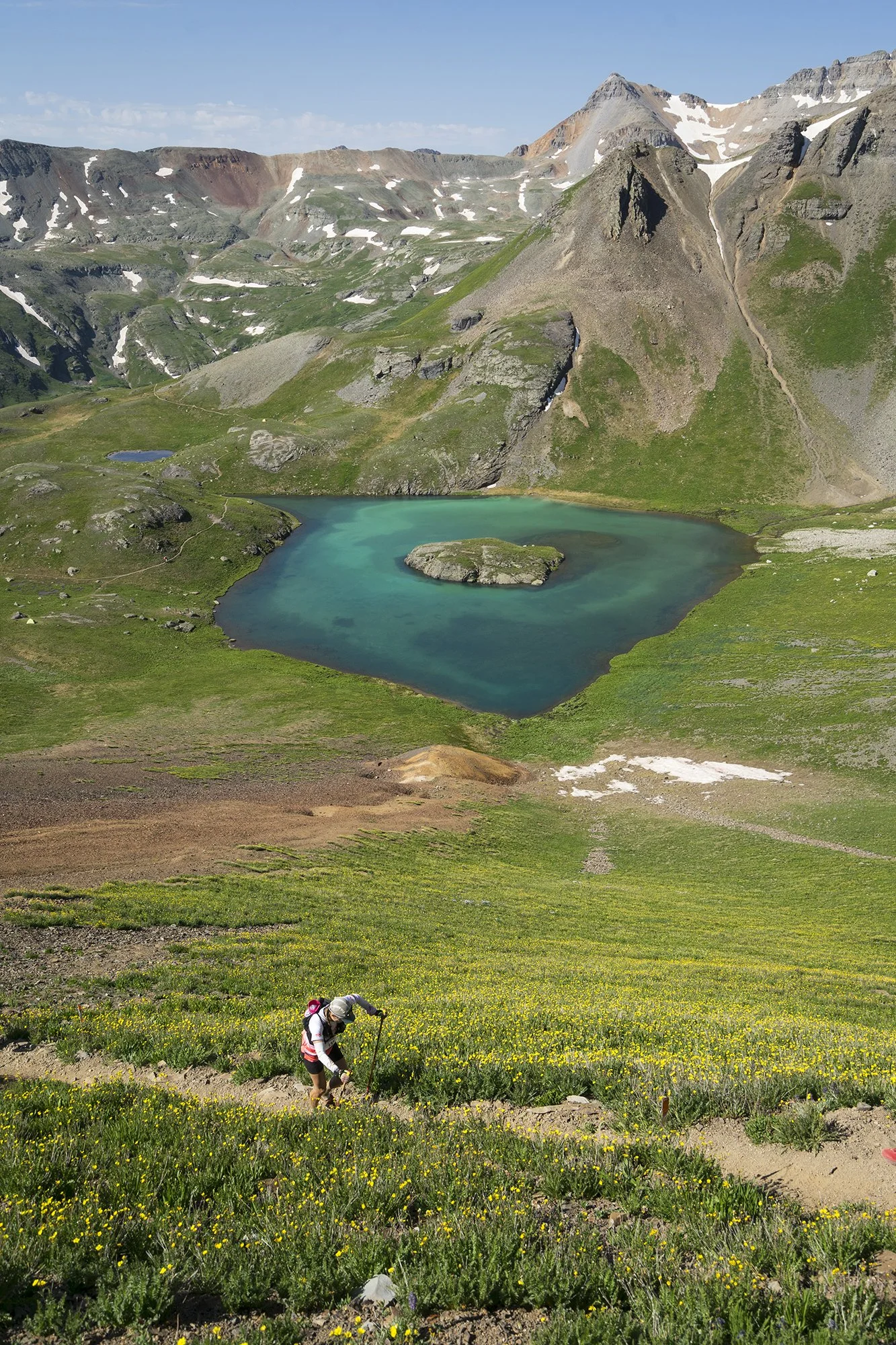 A Hardrock Hundred runner climbs through wildflowers above a turquoise alpine lake near Silverton.